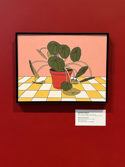 Framed canvas print of a potted plant on a yellow checkered surface with a pink background. Frame is hanging on a red wall.